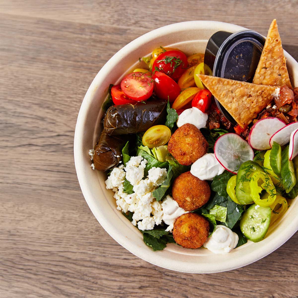 A vibrant Mediterranean-style bowl filled with falafel balls, grape leaves, cherry tomatoes, cucumber slices, radish slices, crumbled feta cheese, pita chips, and a small container of dark dipping sauce, all arranged on a wooden surface.
