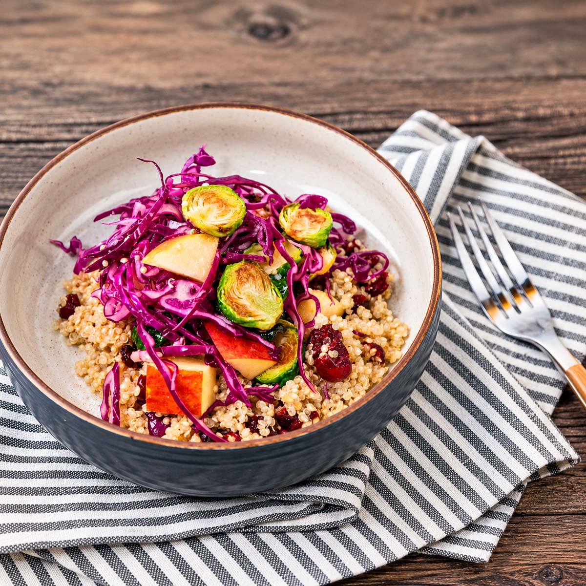 A bowl of colorful quinoa salad topped with sliced Brussels sprouts, red cabbage, and other vegetables, placed on a striped cloth with a fork beside it.