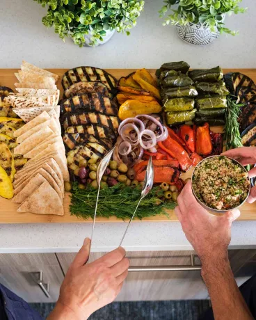 Two people serving themselves from a large wooden platter filled with grilled vegetables, pita bread, olives, stuffed grape leaves, and cheese in a bright kitchen setting.