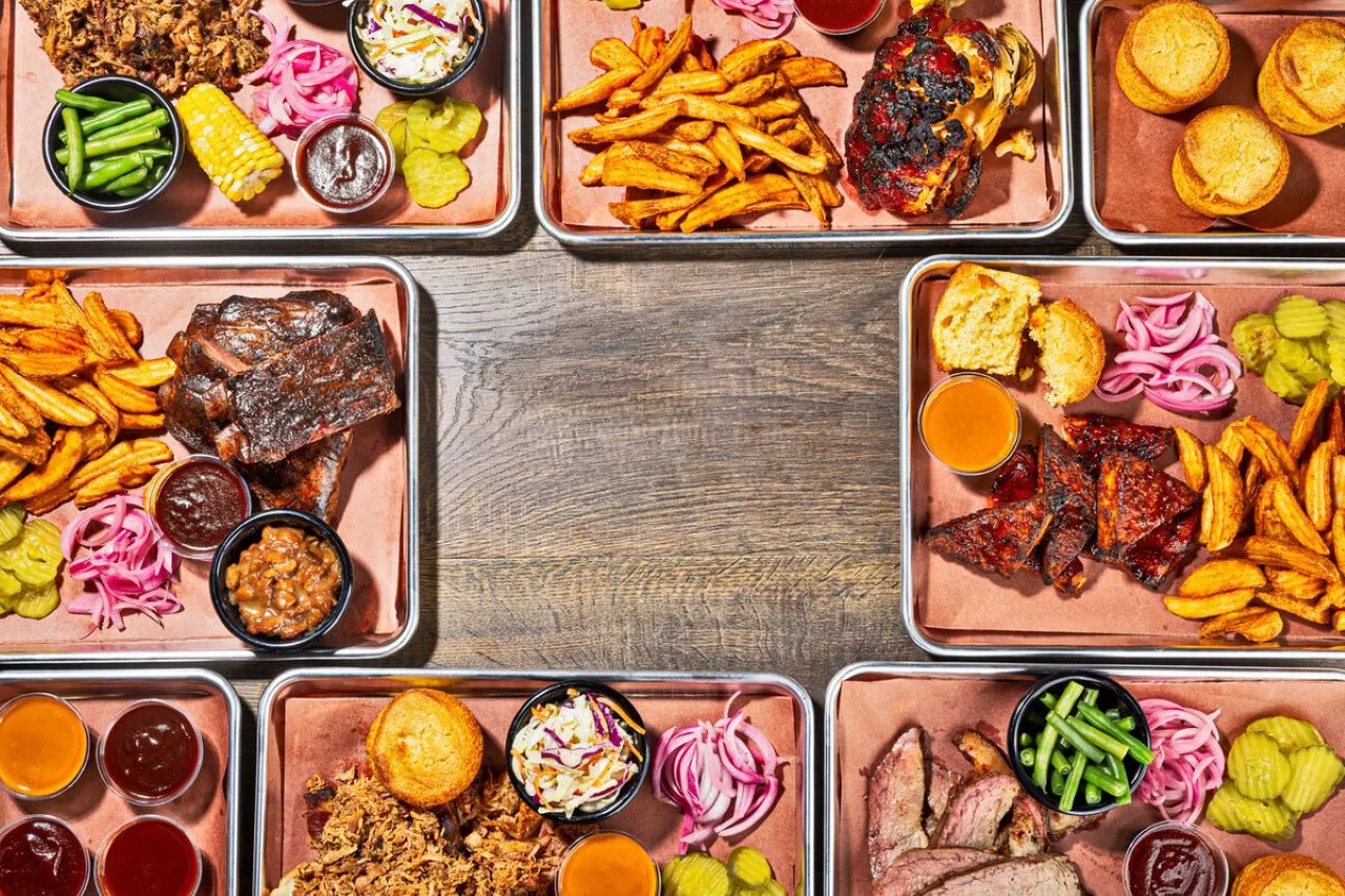 Overhead view of various bbq meat platters with sites on metal trays set on wooden table