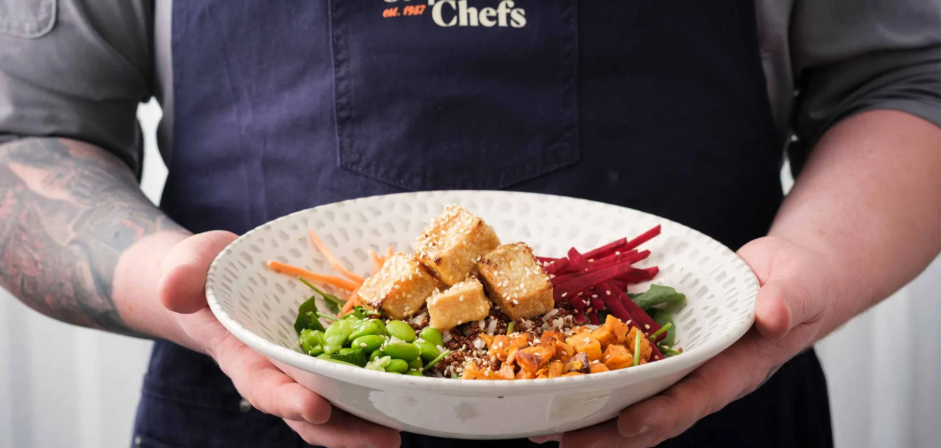 A person wearing a "Corporate Chefs" apron is holding a plate of food featuring tofu cubes, assorted vegetables, and grains in a kitchen or dining setting.