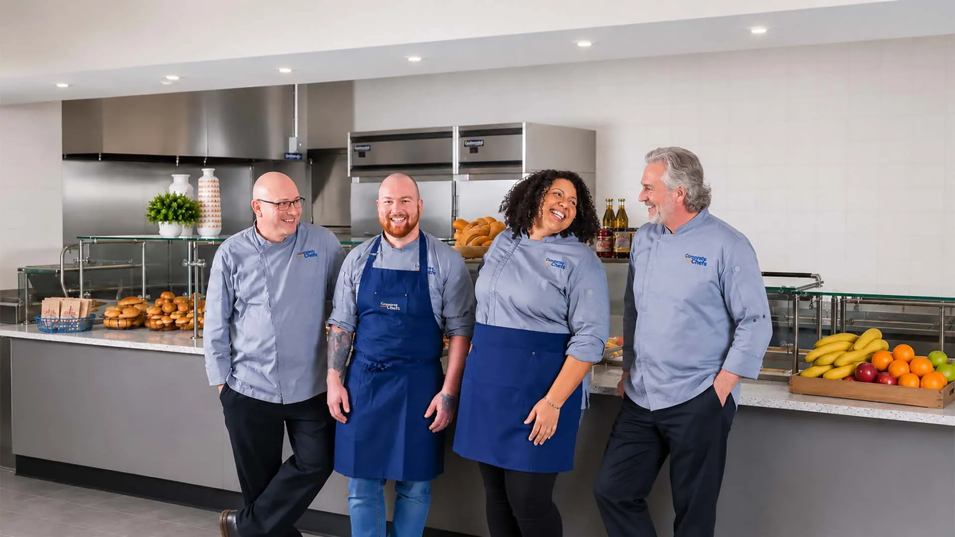 Four chefs standing together in a commercial kitchen, smiling and engaging warmly with each other.