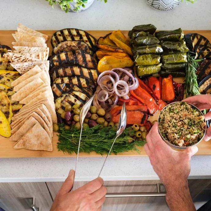 Two people serving themselves from a large wooden platter filled with grilled vegetables, pita bread, olives, stuffed grape leaves, and cheese in a bright kitchen setting.